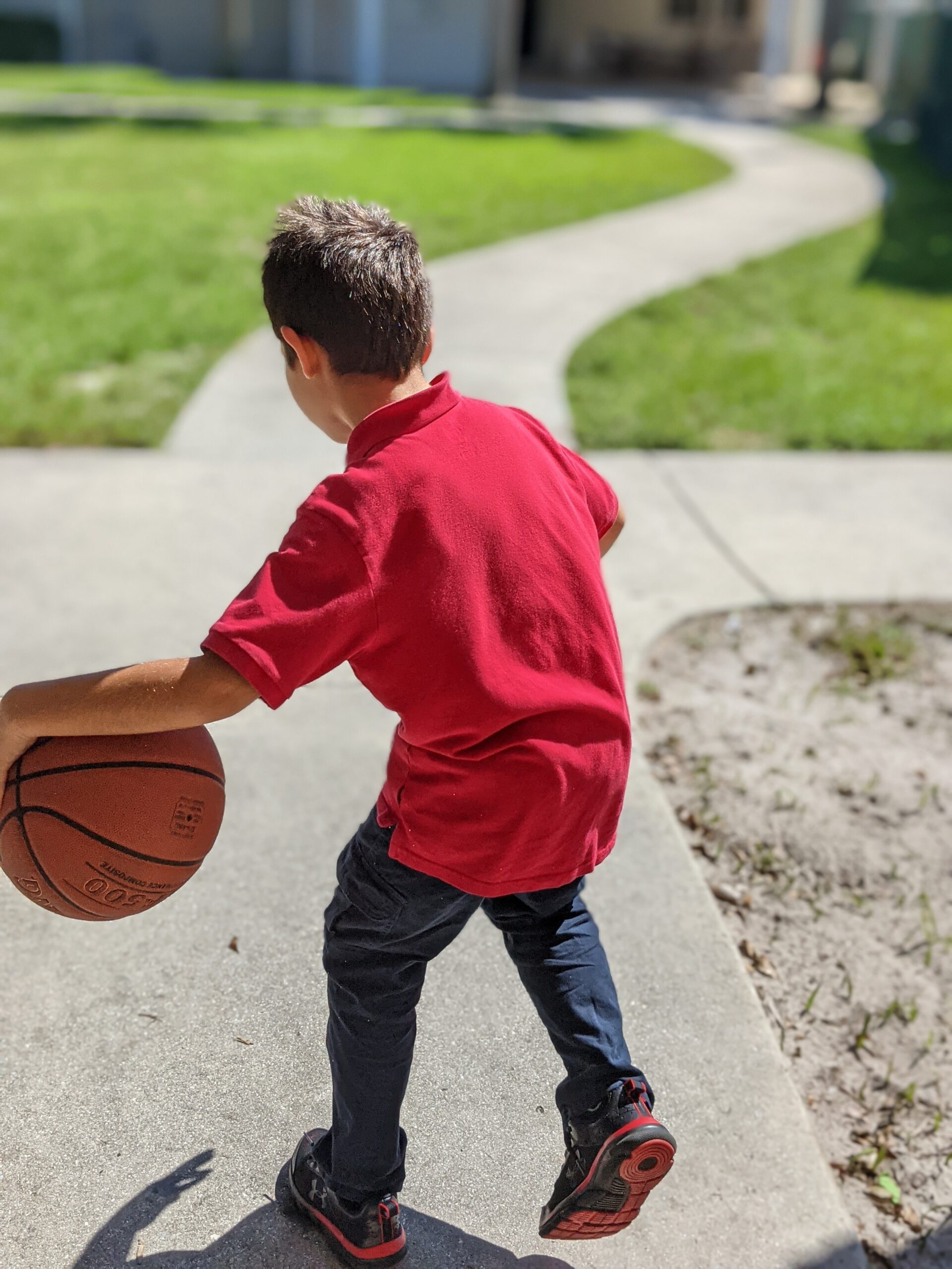 Young boy in red shirt dribbling basketball on sidewalk.